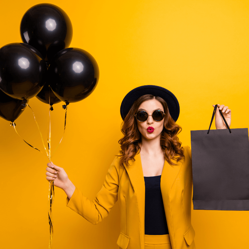 Eine Frau mit Sonnenbrille und Hut hält schwarze Luftballons und eine schwarze Einkaufstüte in den Händen - Titelbild zu "Der ultimative Black Friday-Survival Guide" / Foto von © deagreez via Adobe Stock