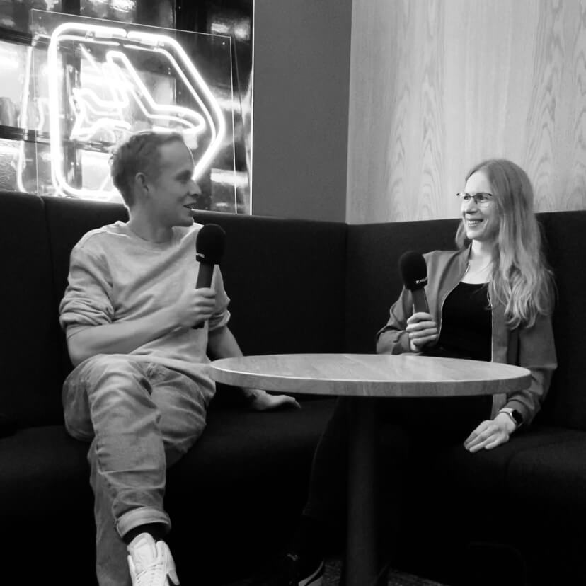 Moritz sitzt mit Hanna Huber auf einer Eckbank, er links, sie rechts, sie halten Mikrofone in der Hand und lächeln