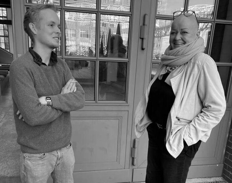 Moritz links, schaut in die Ferne mit verschränkten Armen, Sabine Fischer rechts schaut lachend in die Kamera, beide stehen vor dem Amplifier Berlin