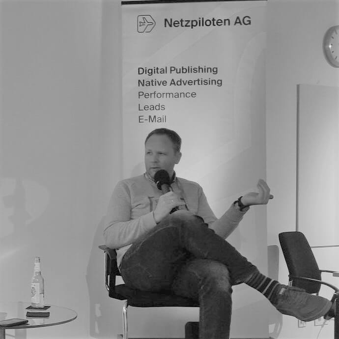 Clas Beese auf der Bühne der FinTech Week mit Mikro in der Hand. im Hintergrund ein Banner der Netzpiloten, er sitzt auf einem Stuhl mit Mikro in der Hand