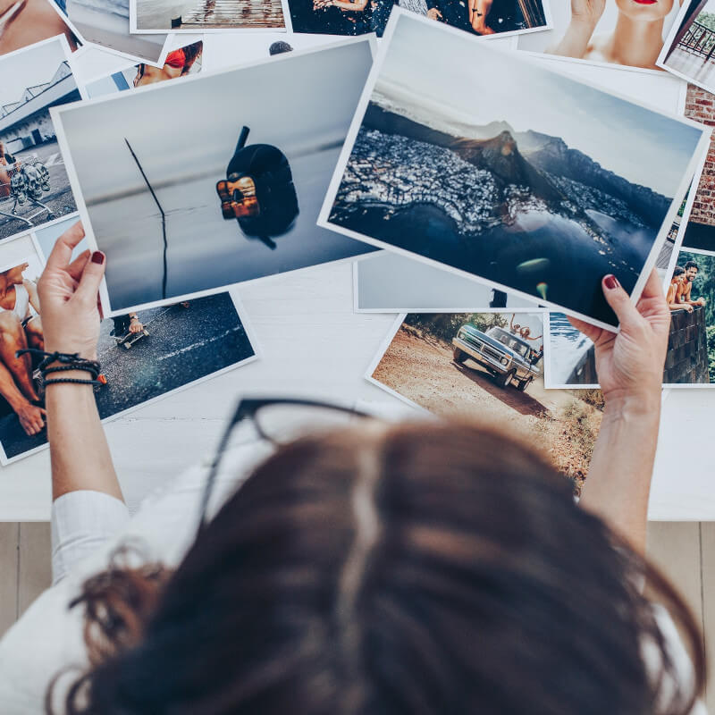Eine Frau sichtet mehrere Fotos. Zwei hält sie in der Hand, viele weitere sind auf dem Tisch verteilt.