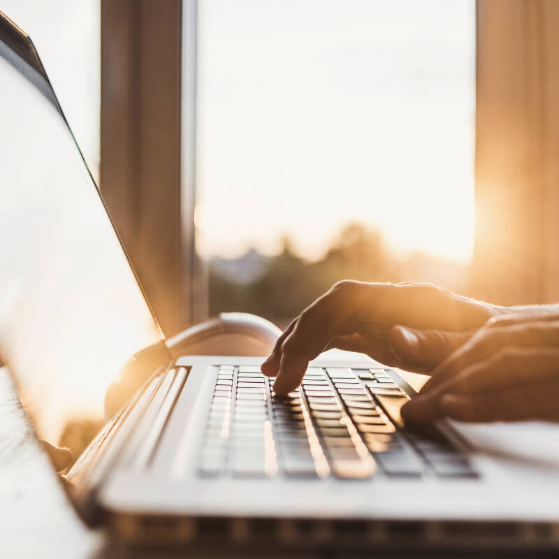 Eine Hand tippt auf einem Laptop, im Hintergrund scheint die Sonne der aufgehenden Sonne durch Fenster.