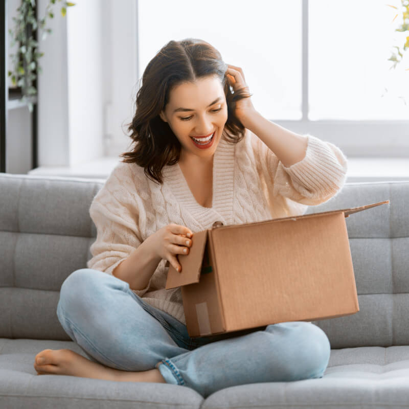 Eine Frau sitzt im Schneidersitz auf dem Sofa und schaut freudig in ein Paket, dass sie gerade geöffnet hat.