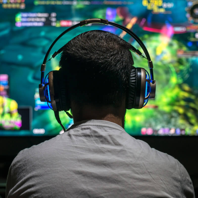 Man sieht einen jungen Mann von hinten, der vor einem Monitor sitzt. Er trägt ein Gaming-Headset mit leichter Beleuchtung.