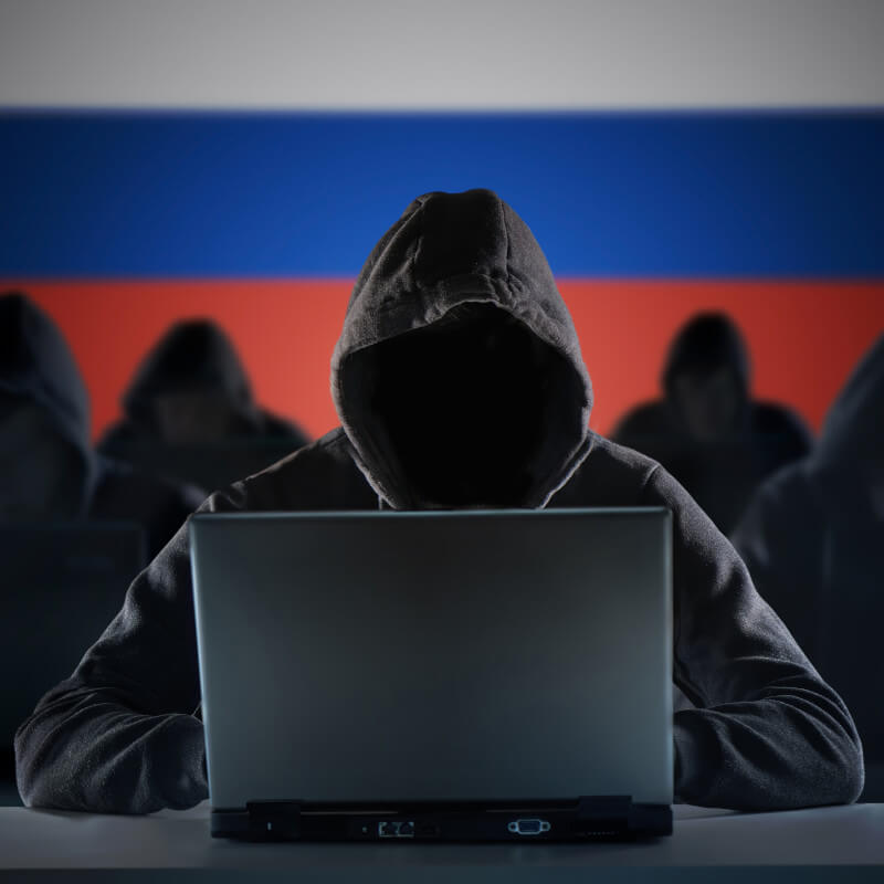 Eine Gruppe unkenntlicher Personen in Kapuzenhoodies arbeiten an Laptops. Der Hintergrund ist eine russische Flagge.