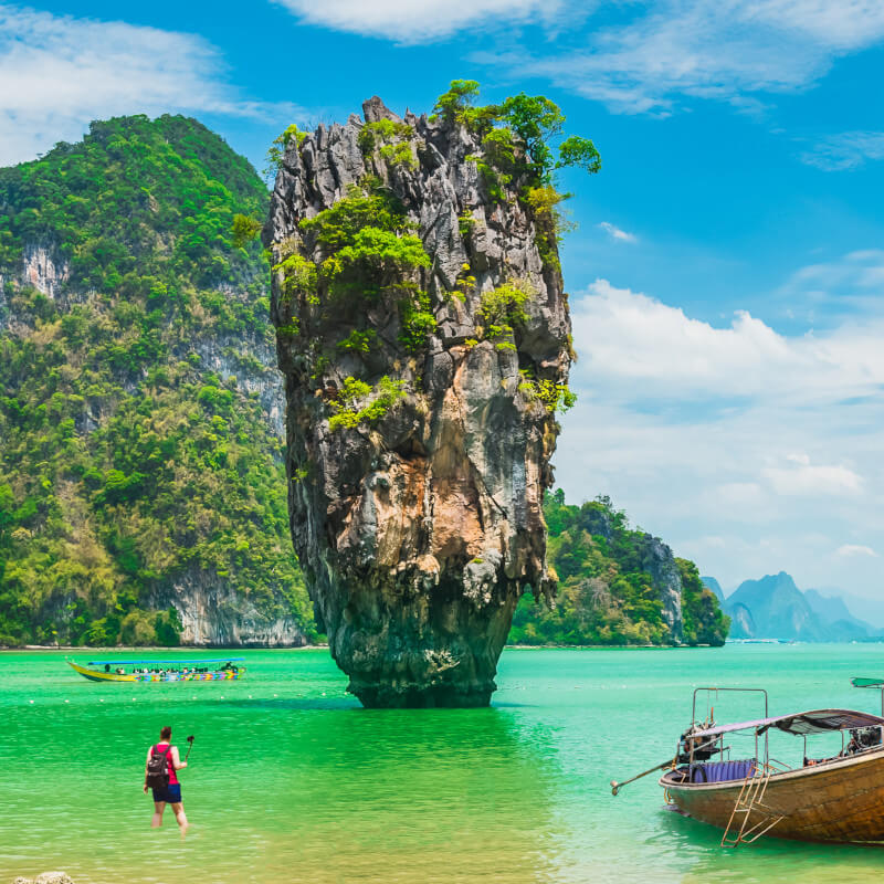 Reisender mit Selfiestick knietief im Meer in Thailand. Im Hintergrund atmenberaubende Berge. Handelt es sich um einen Travel-Vlogger?