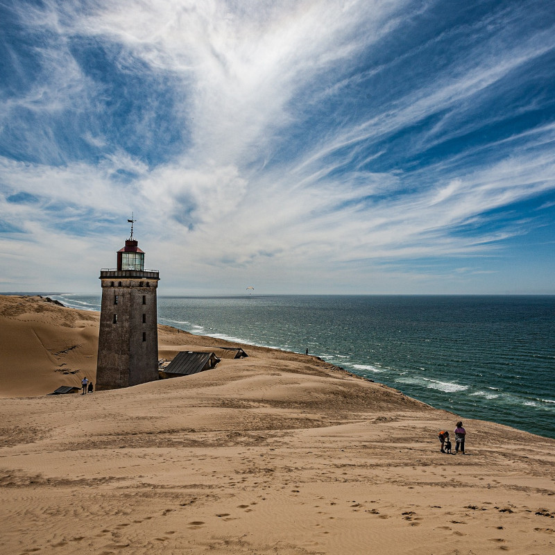 Ein schöner Sandstrand in Dänemark bei schönem Wetter mit einem Leuchtturm und nur vereinzelten Personen, die am Strand entlang wandern.