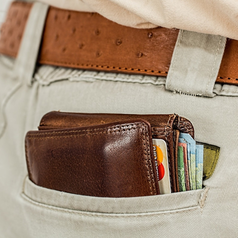 Ein braunes Lederportemonnaie mit Kreditkarten und Bargeld steckt in der hinteren Tasche einer beigen Hose, die mit einem braunen Ledergürtel getragen wird.