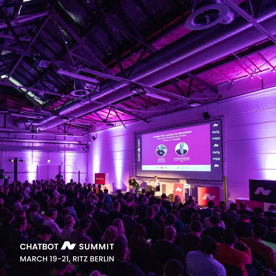 Blick auf eine von violettem Licht beleuchtete Bühne des Chatbot Summit, auf der ein Fireside-Chat vor großen Publikum stattfindet.
