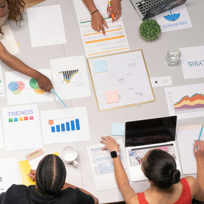 Alt-Text 3: Vogelperspektive auf einen Arbeitstisch, an dem vier Personen sitzen und sich über Marketingmaterialien beugen, die auf dem Tisch ausgebreitet sind. Zu den Materialien gehören Diagramme, Graphen und strategische Dokumente, die auf eine Planungssitzung oder eine Marketinganalyse hinweisen.
