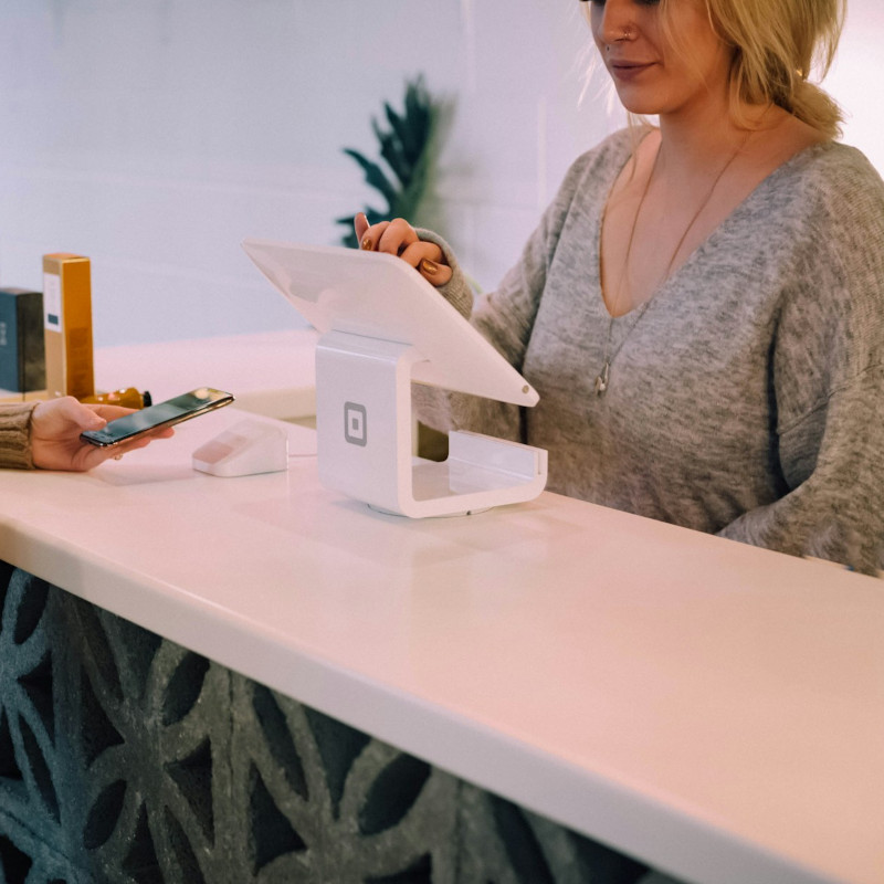 Zwei Frauen stehen sich an einem modernen weißen Tresen gegenüber. Die Frau auf der einen Seite des Tresens hält ein Smartphone in der Hand und ist im Gespräch mit der anderen Frau, die neben einer weißen Point-of-Sale-Station steht. Die Umgebung wirkt einladend und modern.
