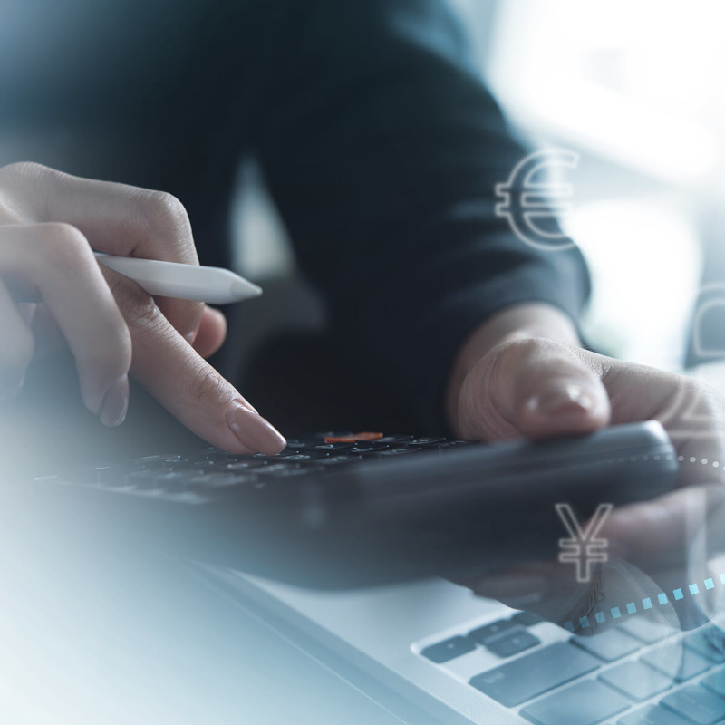 Ein professioneller Arbeitsbereich mit Fokus auf Finanzanalyse. Im Vordergrund tippt eine Person mit einem Stift in der Hand auf einem Taschenrechner, wobei ihre Hände scharf im Fokus sind. Im Hintergrund verschwimmen visuelle Darstellungen von Währungssymbolen und Aufwärtstrends auf Diagrammen, die die globale Finanzwirtschaft symbolisieren.