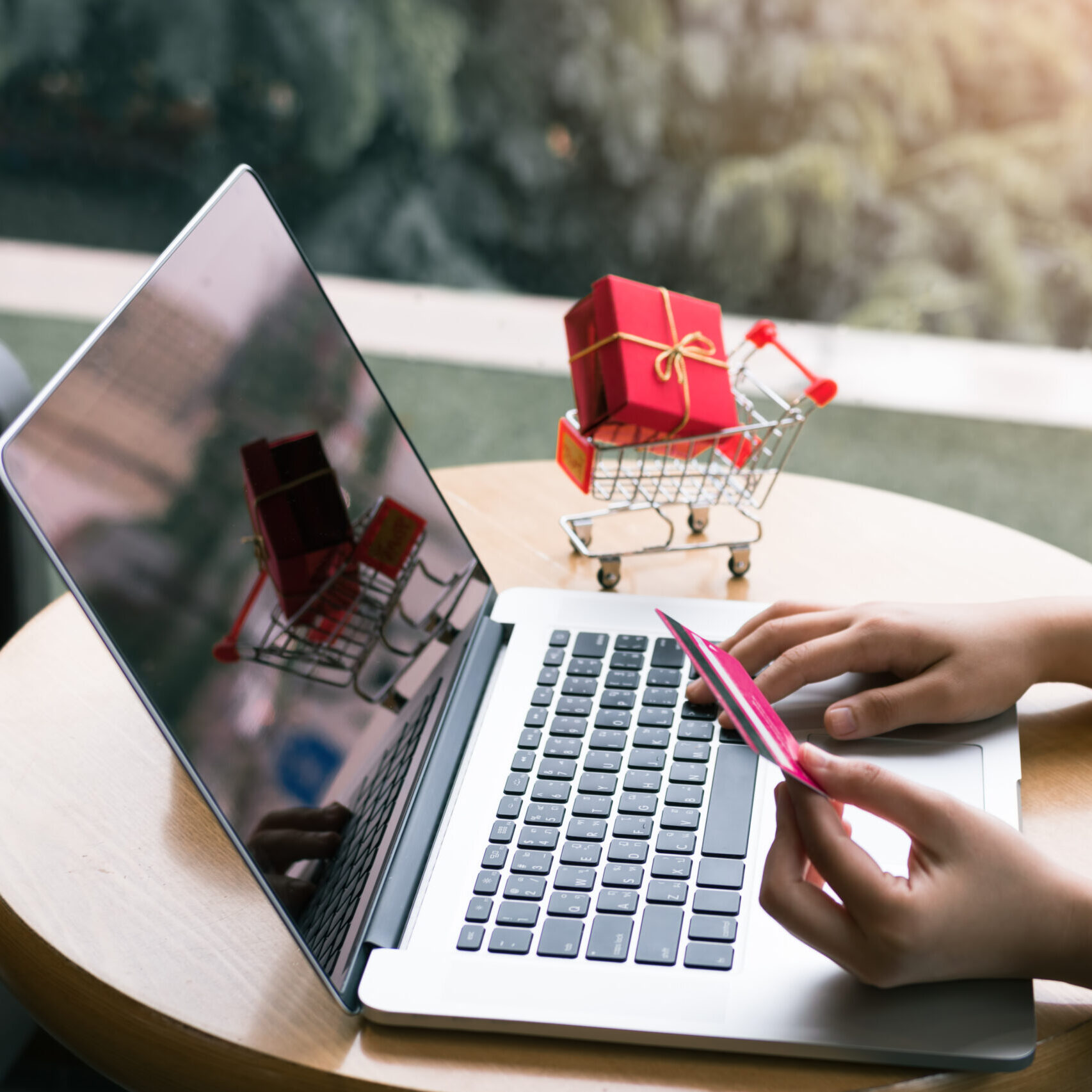 Symbolbild für PSN Gutscheinkarten, Person sitzt an LAptop und hält eine Bezahlkarte in der Hand, im Hintergrund ein Einkaufswagen mit einem verpackten GEschenk darin