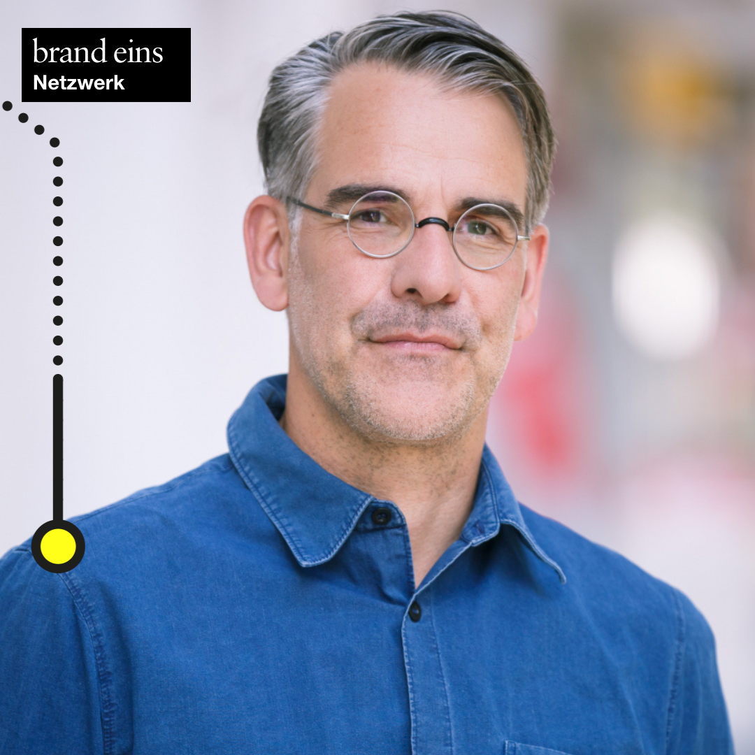 Das Bild zeigt einen Mann mittleren Alters mit graumelierten Haaren und einem gepflegten Bart. Er trägt eine runde Brille und ein blaues Hemd. Der Hintergrund ist verschwommen, wodurch der Fokus auf dem Mann liegt. In der oberen linken Ecke befindet sich ein Logo mit der Aufschrift "brand eins Netzwerk".