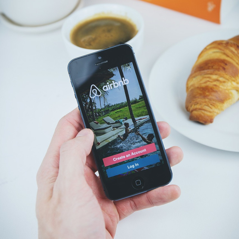 Ein Smartphone mit geöffneter Airbnb-App wird in der Hand gehalten. Im Hintergrund befinden sich eine Tasse Kaffee, ein Croissant auf einem weißen Teller sowie Bücher und eine Pflanze. Diese gemütliche Szene erinnert an Reisevorbereitungen und die Suche nach einer Unterkunft - möglicherweise sogar nach den vier besten Airbnb-Alternativen.