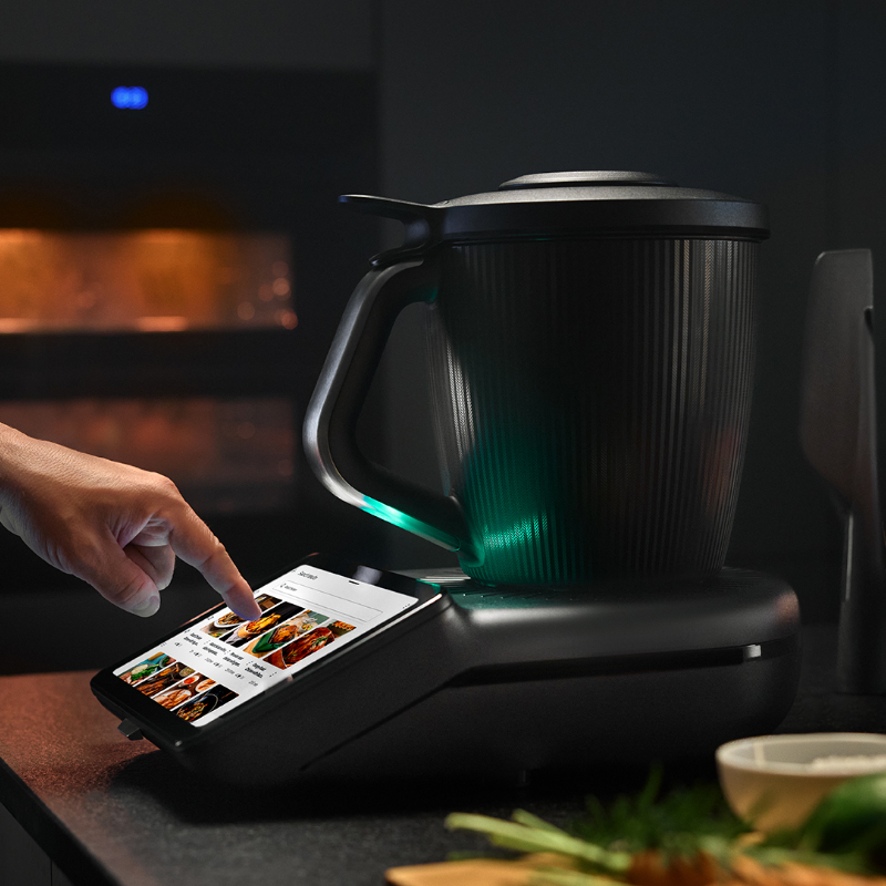 Der neue Thermomix TM7 mit großem Touchscreen: Eine Person tippt auf das Display eines schwarz-matten Thermomix TM7, während der beleuchtete Mixtopf auf der Basis steht. Im Hintergrund ein Backofen und frische Zutaten auf der Arbeitsplatte.