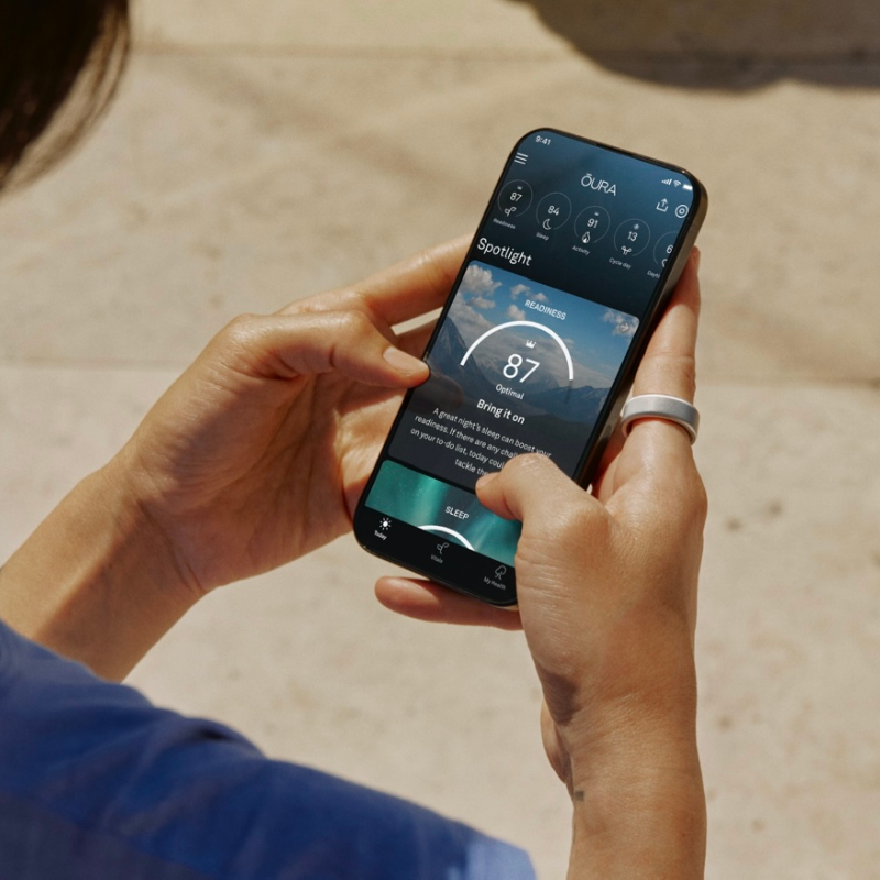 Ein Mensch hält ein Smartphone in beiden Händen und nutzt eine Gesundheits-Tracking-App. Am rechten Zeigefinger trägt die Person einen silbernen Smart Ring. Die App auf dem Bildschirm zeigt eine "Readiness"-Bewertung von 87 an, begleitet von weiteren Gesundheitsmetriken. Der Hintergrund ist leicht verschwommen und zeigt einen sonnigen Außenbereich mit einem hellen Bodenbelag. Die Person trägt ein blaues Oberteil.
