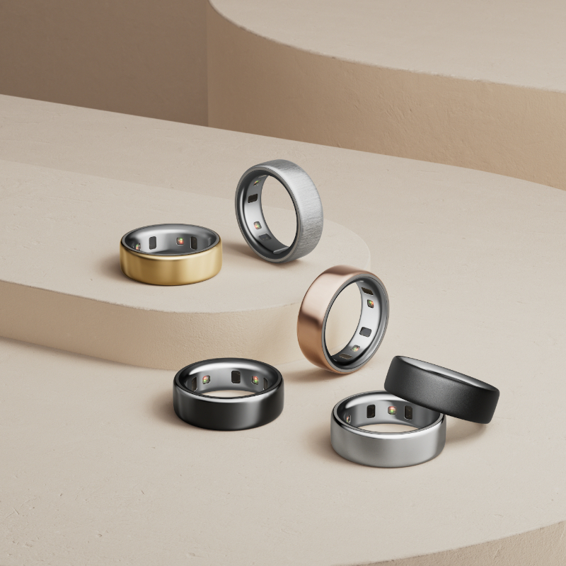 Ein stilvoll arrangiertes Set aus sechs modernen Smart-Ringen in verschiedenen Metallfarben – darunter Gold, Schwarz, Silber, Roségold und gebürstetes Grau – auf einer minimalistischen beigen Stufenplattform. Die Ringe sind mit eingebauten Sensoren auf der Innenseite ausgestattet und reflektieren einen eleganten, technologischen Look. Die Komposition betont die luxuriöse Verarbeitung und die Vielseitigkeit der Smart-Ringe als modische Wearables.