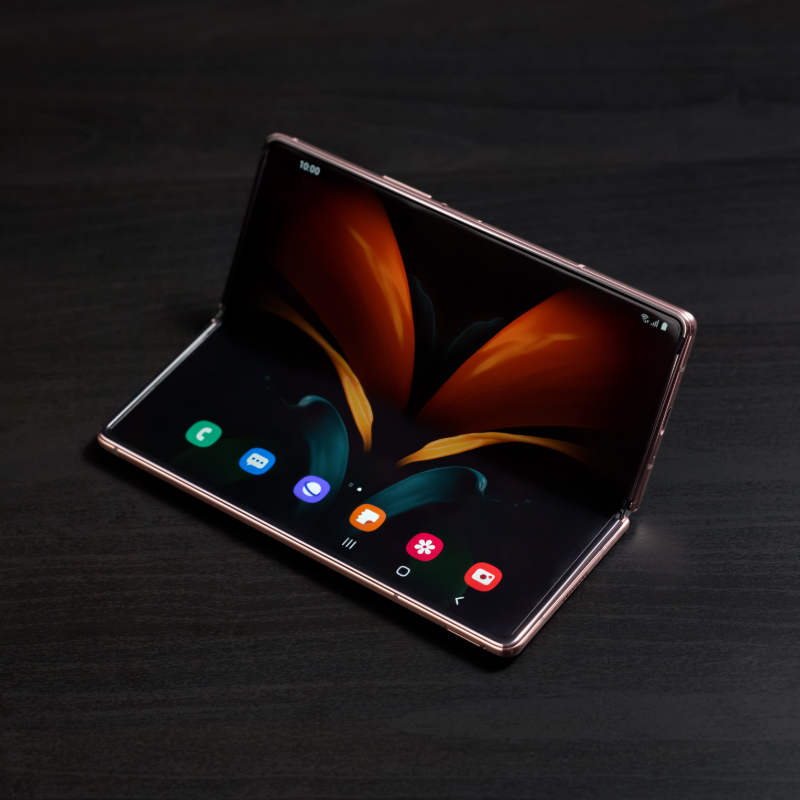 Ein modernes faltbares Smartphone liegt auf einer dunklen Oberfläche und ist halb aufgeklappt. Der große, flexible Bildschirm zeigt eine elegante, abstrakte Hintergrundgrafik mit leuchtenden Farben. Die Benutzeroberfläche ist sichtbar, mit mehreren App-Symbolen am unteren Rand des Displays. Das Gerät hat ein schlankes, hochwertiges Design mit metallischem Rahmen und nahtlosem Displayübergang.