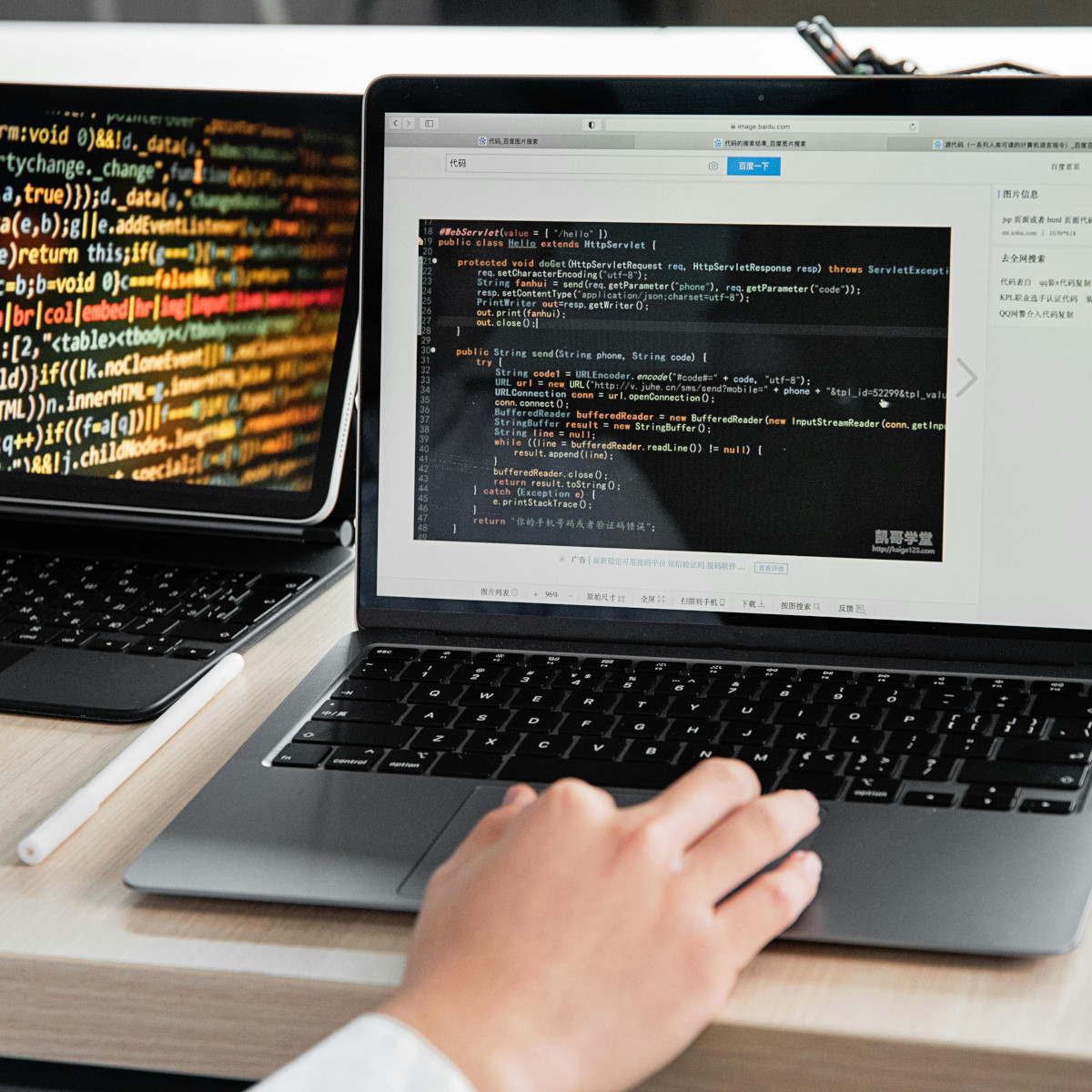 Auf einem Tisch stehen ein Tablet und ein Laptop. Auf den Bildschirmen ist Code zu sehen und eine Hand beient grade die Tastatur des Laptops.