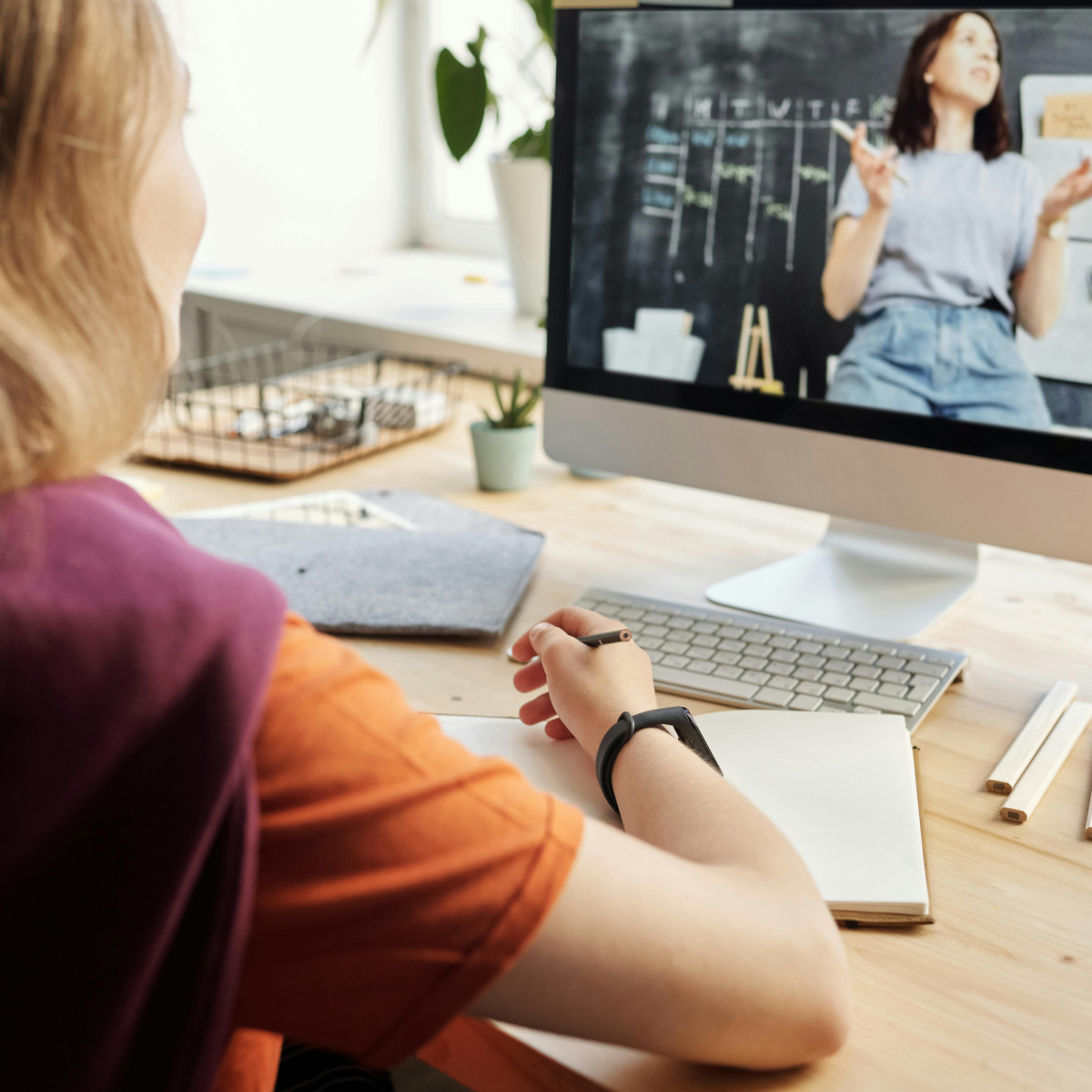 Eine Frau sitzt mit Stift und Heft vor ihrem Computerbildschirm. Auf dem Bildschirm ist eine weitere Frau, eine Lehrperson, vor einer Tafel zu sehen.
