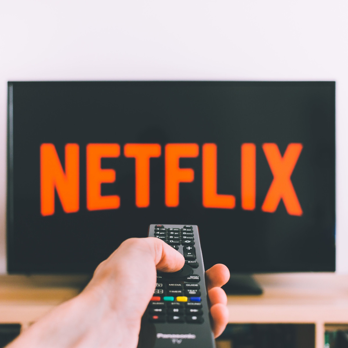Im Hintergrund ist ein Fernseher mit dem Netflix-Logo, davor richtet eine Hand eine Fernbedienung darauf